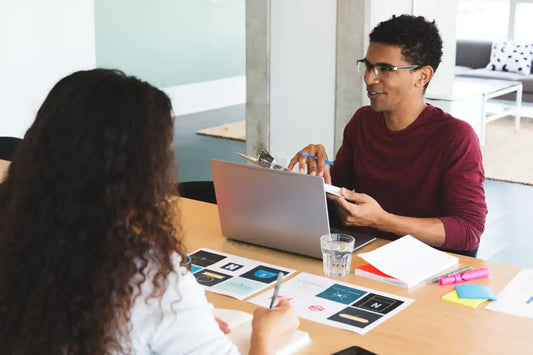 Two people sitting at a desk with a laptop and office supplies, engaged in conversation or a meeting.